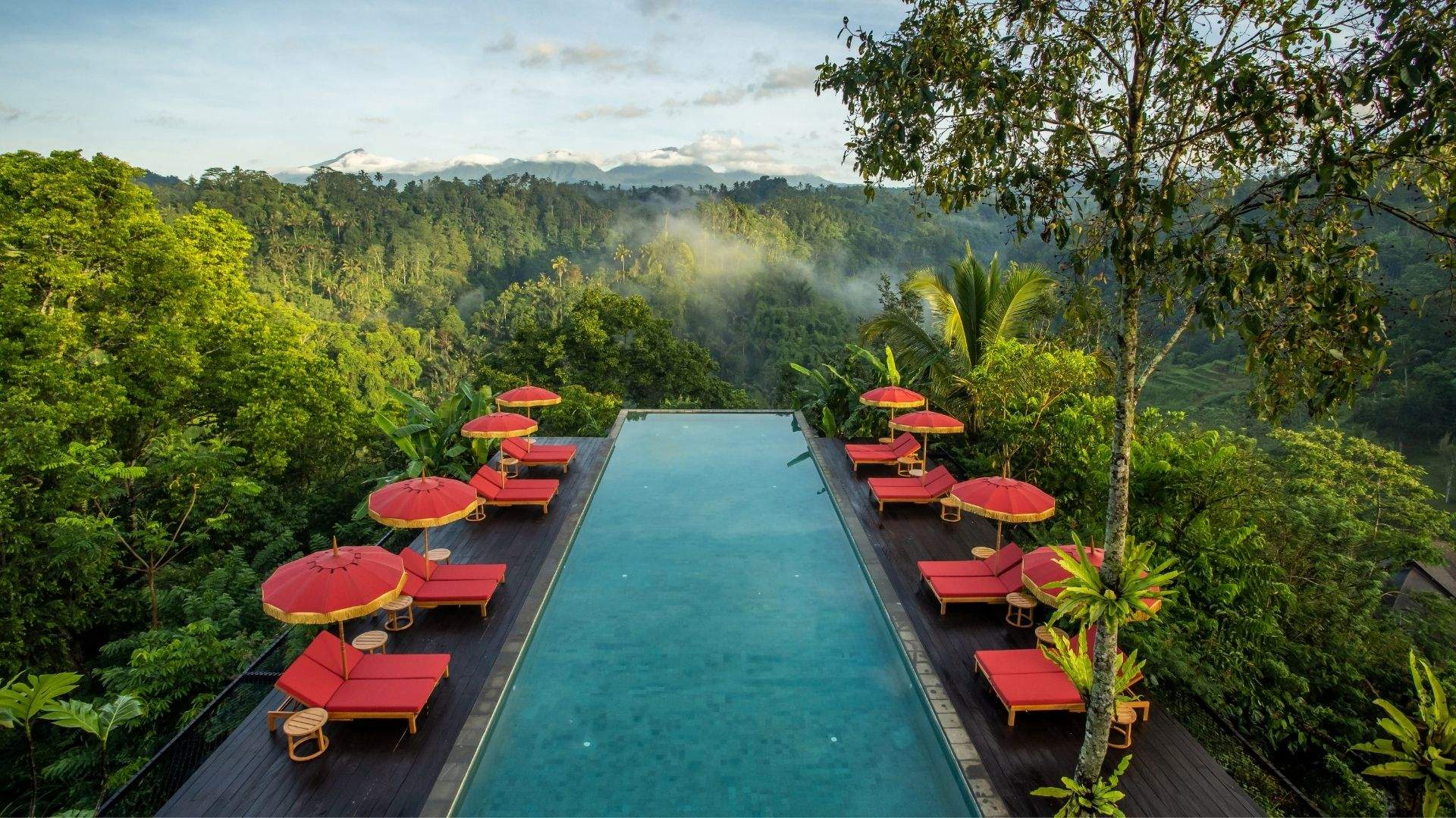 Infinity pool reflects sky, lined with red loungers, amid lush forest at Banyan Tree Escape - Buahan.