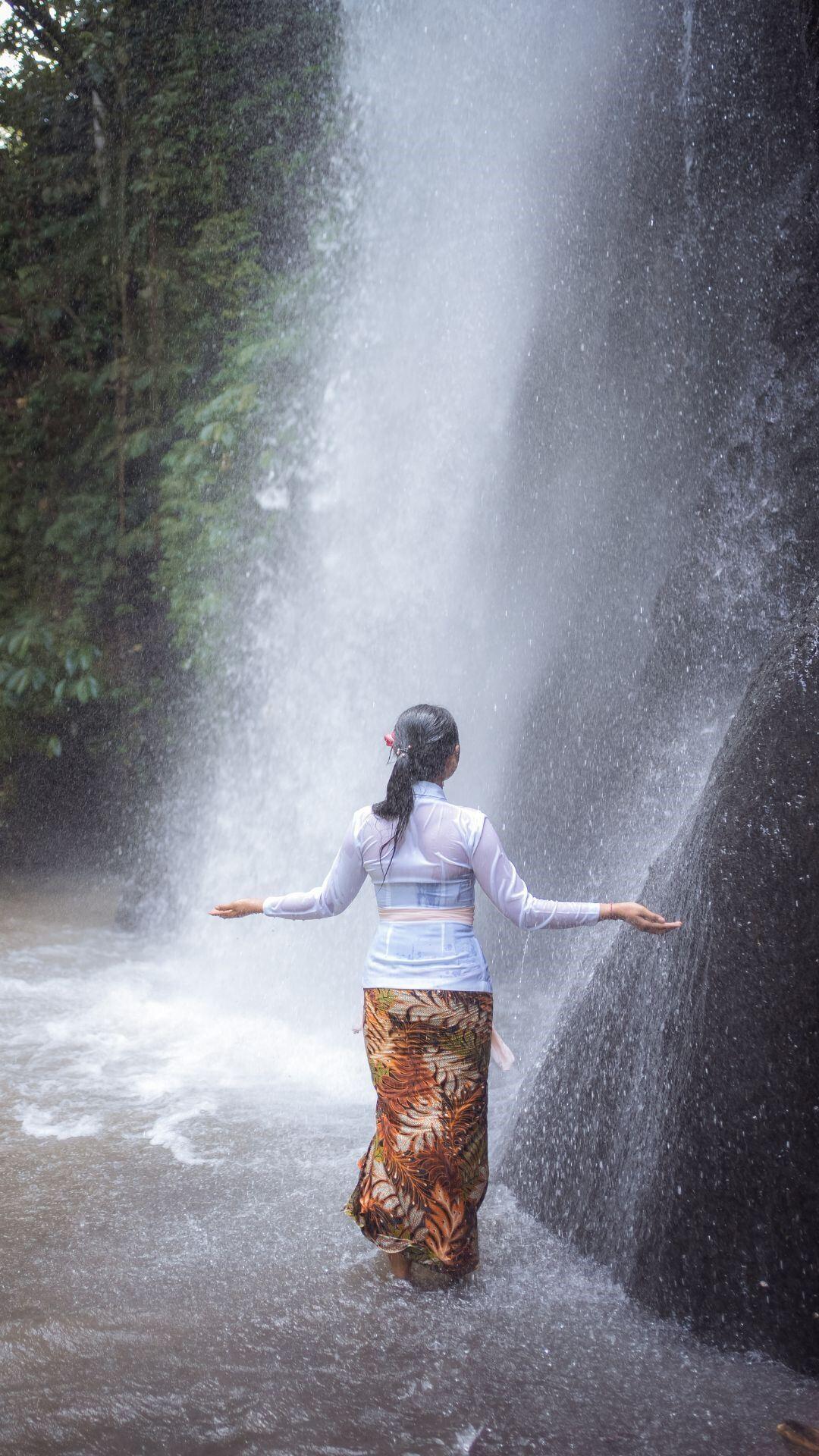 Person stands under waterfall, arms outstretched, surrounded by lush greenery at Banyan Tree Escape - Buahan.