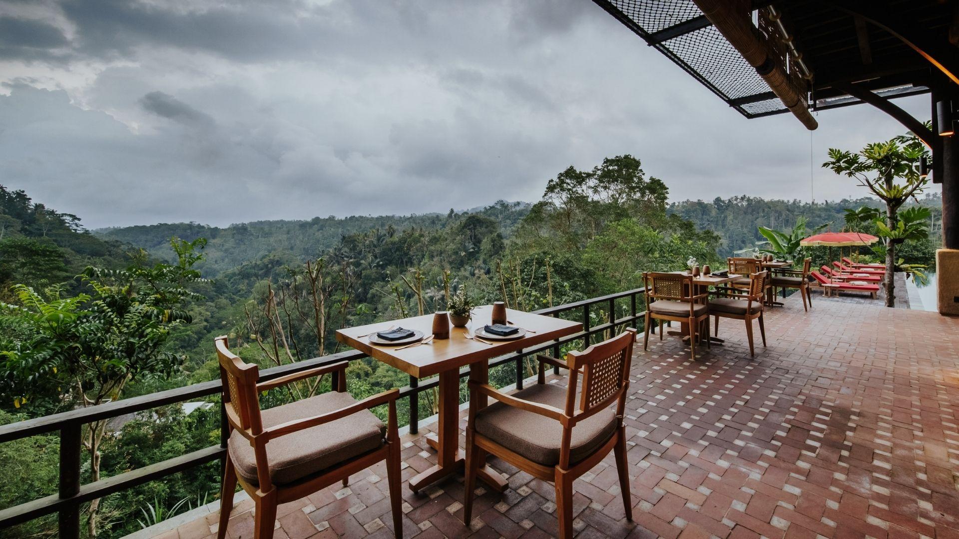 Terrace dining set of Open Kitchen Restaurant, overlooking lush rainforest at Banyan Tree Escape - Buahan, under cloudy skies.