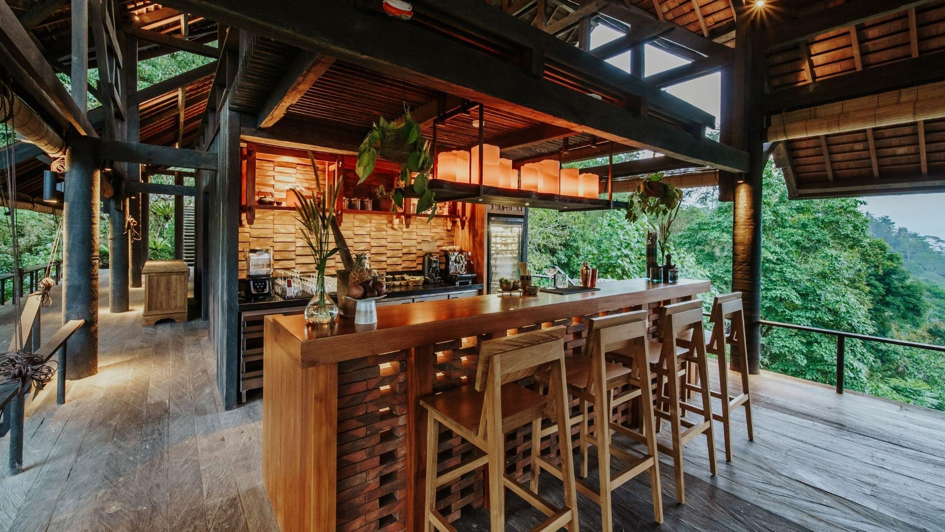 The Botanist bar with wooden stools and hanging lights, nestled in lush greenery at Banyan Tree Escape - Buahan.