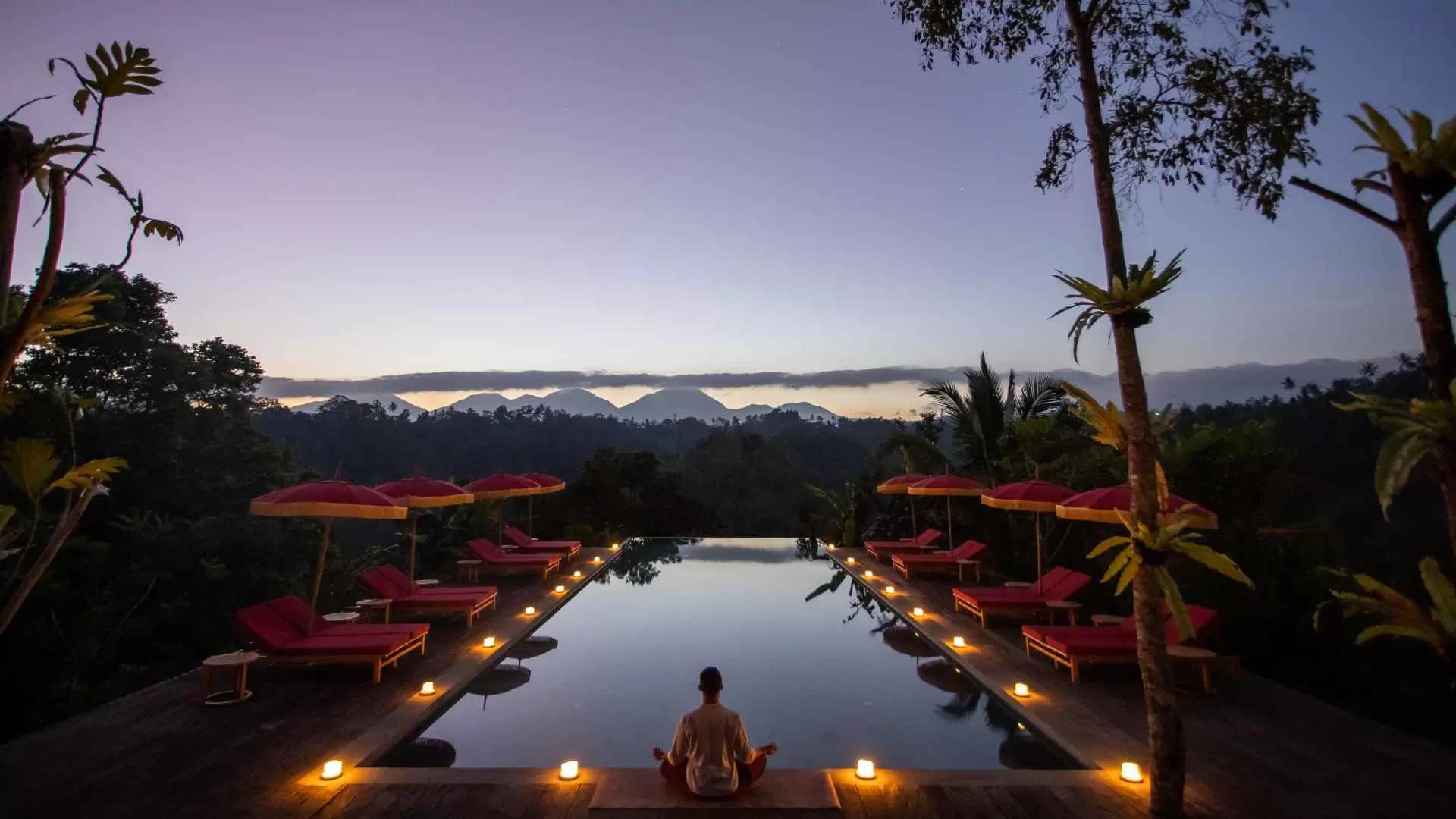 Person meditating of moon yoga by a candlelit pool at Banyan Tree Escape - Buahan, amidst a mountain backdrop.